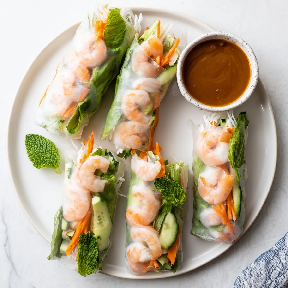 Overhead view of vibrant Vietnamese Spring Rolls on a white plate, with mint leaves, lime wedges, and a rich peanut dipping sauce.